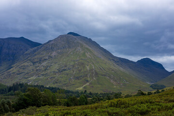 The stunning hills of Glen Coe on a late summer day with moody overcast sky, lush green trees and empty roads.