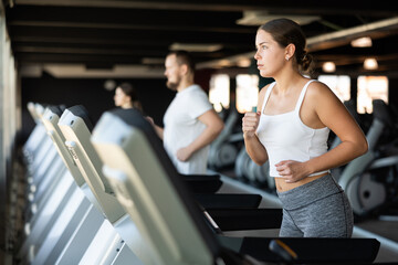 Against background of dark gym of fitness club, visitors and girl coach are clearly visible. She...