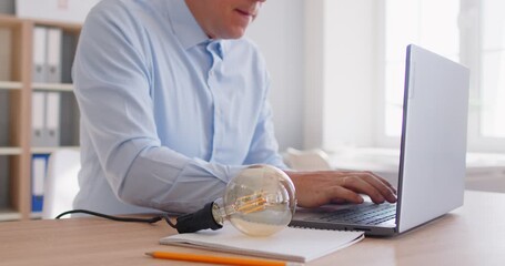 Technology, creativity. Light bulb next to businessman instantly lights up, symbolizing moment of inspiration or new idea. Man in office is typing on laptop and raising his finger up to show new idea. - Powered by Adobe