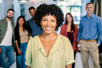Confident young African American woman smiling in office with diverse team in background. Leader standing proudly in a modern workspace. Positivity and professional empowerment concept.