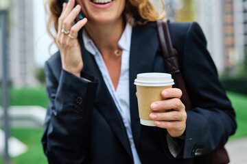 Portrait of a young businesswoman woman using a smartphone mobile phone and holding a cup of coffee walking down the street, surrounded by moder corporate office buildings