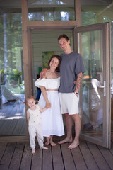 Fototapeta premium A joyful family stands on the porch of their cabin, smiling happily. The parents are dressed casually, and their child is playfully holding hands, surrounded by nature