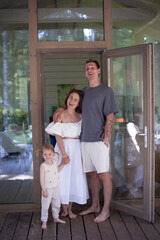 A family stands on a wooden porch of their summer home, surrounded by greenery. The parents look happy while their young daughter holds her mother's hand, creating a joyful moment © Yana Gavriloski