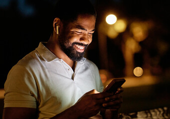 Focused man using smartphone and listening to music song or a podcast outdoors during twilight with city lights in the background