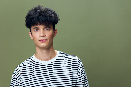 young man with curly hair wearing a striped t-shirt posing against a plain muted green background with a calm and neutral expression
