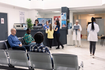 Busy hospital lobby with patients and visitors seated in waiting area. Receptionist and nurse...
