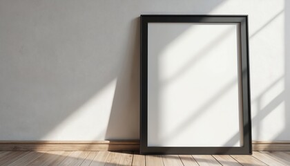 Empty black photo frame leans against textured wall, white matting borders blank space. Wooden floorboards show diagonal light patterns from window, sunbeams cross frame surface. Clean indoor
