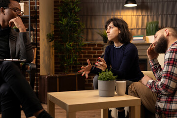 Therapy session with African American psychiatrist mediating conflict between caucasian couple. Frustrated wife shows smartphone while stressed husband rubs head, tension in relationship counseling.