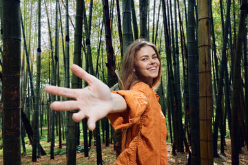 Naklejka premium Woman smile bamboo forest hand reaching toward camera, portrait of happy young woman outdoors in nature wearing orange jacket, looking back and inviting viewer on a woodland walk.