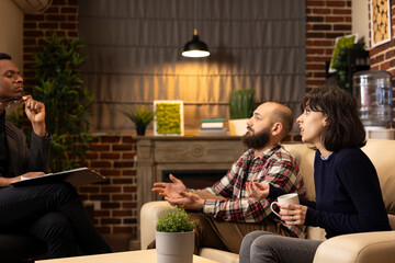 Psychiatrist sits with clipboard on knee while caucasian couple discusses relationship conflicts on couch. Therapist listens attentively, analyzing marriage issues and providing professional guidance.