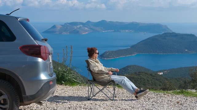 Woman relaxing in folding chair by car overlooking picturesque seascape. Tourist enjoying breathtaking ocean view from mountain viewpoint, relaxing in a portable chair next to her vehicle
