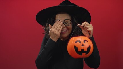 Middle age hispanic woman covers face with hand while holding a pumpkin basket in a studio; festive halloween joy surprise. - Powered by Adobe