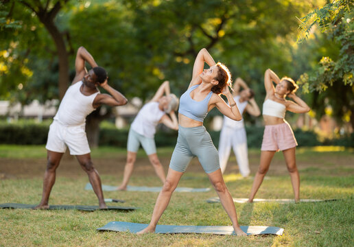 Multinational people of different ages in park practicing yoga. Amateurs performing gymnastic. Woman yoga master and followers do Tiryaka Tadasana. People practice during sport in nature