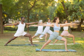 During street workout, yoga practice on lawn in public park, multinational athletes of different ages fulfil carry out warrior pose virabhadrasana exercise. Students attend outdoor yoga lesson in park