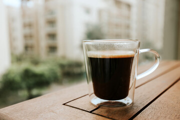Freshly brewed coffee in a glass cup on a wooden table with a blurred city background