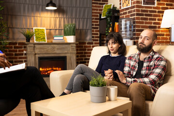 Portrait of caucasian spouses seated on couch in brick wall office, smiling with relief after therapy session. Husband and wife look at camera during positive counseling discussion with therapist.