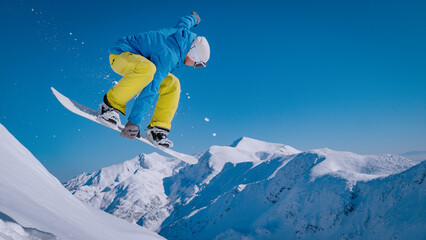 CLOSE UP, LOW ANGLE VIEW: Snowboarder launches off a steep ridge and flies above scenic snowy...