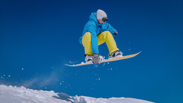 CLOSE UP, LOW ANGLE VIEW: Snowboarder in blue jacket performs indy grab trick while riding off the piste. Fresh powder snow trails beneath the board against deep blue sky. Pure winter action and style