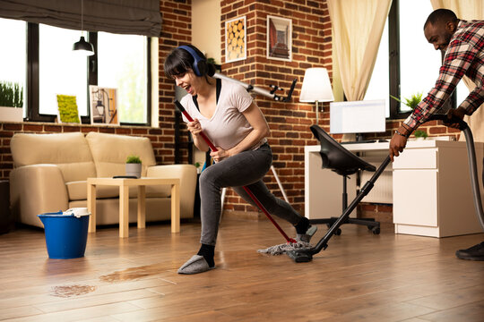 Multiethnic couple enjoys playful spring cleaning in brick wall apartment. Girlfriend with headphones, sings into mop stick as smiling boyfriend vacuums floor, turning chores into fun bonding moments. - Powered by Adobe