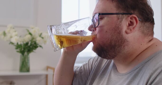Overweight man eating fries at home, glasses on. He slowly drinks beer from a glass, enjoying a casual snack while watching TV. Plus size plump person enjoying unhealthy fast food. 