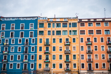 A symmetrical, full-frame view of three adjacent, brightly colored apartment buildings in Bilbao, featuring distinct blue, yellow-ochre, and orange facades with repetitive window and balcony patterns.