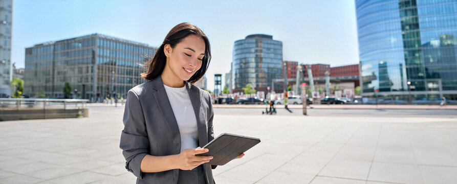 Young happy Asian professional woman wearing suit holding digital tablet standing in big city on busy street, smiling lady using smart business software for online work on pad computer outside