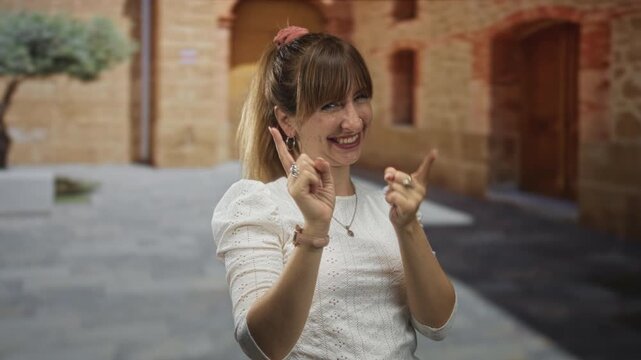 Woman wearing white eyelet blouse and necklace points index fingers while smiling on old town street, near stone archways and tree; playful local charm.