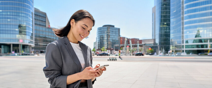 Young happy Asian business woman wearing suit holding mobile phone standing in big city on busy downtown street, smiling lady using smartphone for texting, using apps outside