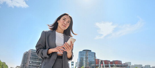 Young Asian businesswoman wearing suit holding mobile phone standing in big city on busy downtown street. Young chinese business woman using smartphone texting, using apps outside