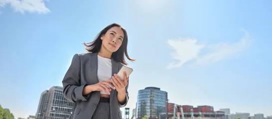 Selbstklebende Fototapeten Sportgeschäft Young Asian businesswoman wearing suit holding mobile phone standing in big city on busy downtown street. Young chinese business woman using smartphone texting, using apps outside  © insta_photos