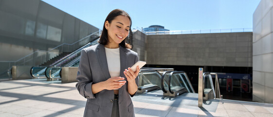 Young happy Asian business woman wearing suit holding mobile phone standing in city subway using...