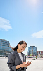 Young happy Asian business woman wearing suit holding mobile phone standing in big city on busy...