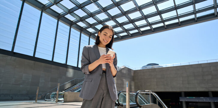 Young smiling Asian business woman wearing suit holding cell phone standing in city metro using smartphone fast connection, checking apps for public transport or travel navigation maps guide