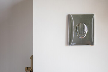 Chrome flush plate on tiled wall, compact actuator detail with reflective surface, neutral grout...