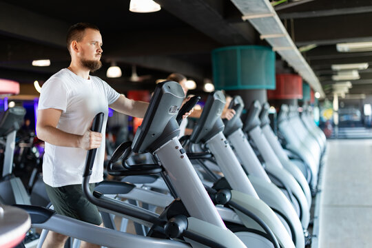 Young man stands on simulator, performs fitness task using an elliptical simulator. Visitor usefully spends time in fitness club, gets body in good physical shape.