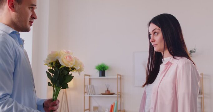 Young guilty man standing in living room with bouquet of flowers apologizing to angry girlfriend after conflict at home. Couple quarreling having marriage problems. Relationships concept. 4k video.