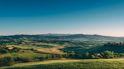 Obraz premium Rolling cultivated hills stretch toward distant mountains under a clear blue sky at sunrise