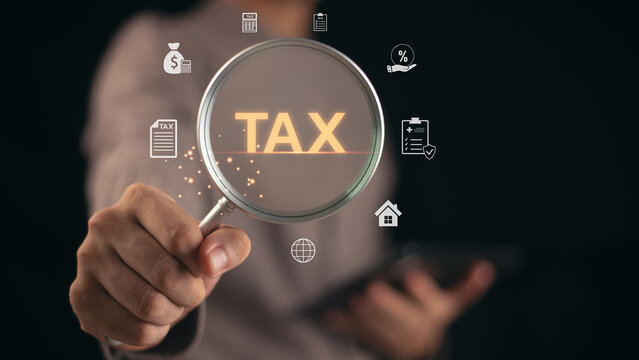 A businessman holding a magnifying glass with the word TAX glowing in focus, representing financial audit, tax calculation, and business accounting concept for digital finance and corporate taxation.