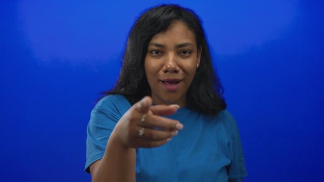 African american woman wearing blue t shirt points finger directly at camera in studio setting; invitation interaction.