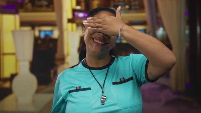 Woman covers eyes with hand in hotel building while holding a whistle and wearing referee uniform; shyness.