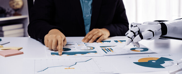 A businessman focuses on analyzing financial reports with the assistance of a robot hand in a sleek modern office, highlighting the integration of technology in business.Noogenesis