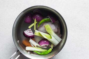 Overhead view of ingredients for making chicken broth in a silver saucepan, top view of raw chicken carrots green chilli spring onions in a saucepan, process of making chicken broth