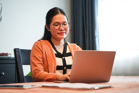 Asian woman working remotely on laptop in cozy home setting, Focused female professional typing on computer at home workspace, Woman using laptop for online work or study in bright living room