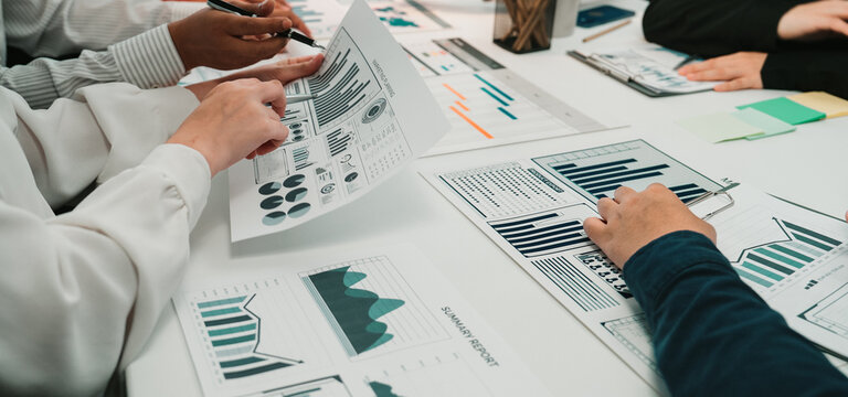 A diverse group of professionals collaborates in a business meeting, analyzing financial reports and data charts spread across a table, showcasing teamwork and engagement. SACTR