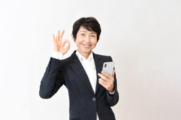スマホを持ちながらOKサインをするスーツ姿のアジア人のミドル女性
