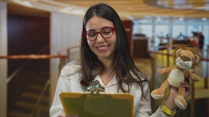 Woman smiling indoors hotel holding clipboard and toy lion wearing red glasses highlighting cheerful young hispanic hospitality professional in casual setting