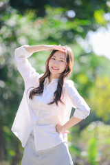 A young beautiful Asian woman in a white shirt and light pants shields her eyes from bright sun outdoors. She is smiling and posing with a green bokeh background.