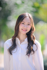 beautiful smiling young Asian woman with long wavy hair wearing a white shirt, posing outdoors in bright sunlight with a blurred green background