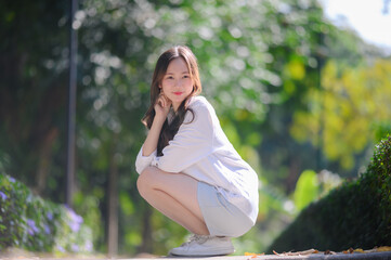 A young beautiful Asian woman in a white shirt and light skirt crouches outdoors on a pathway. She is smiling and looking up with a blurred green park background.
