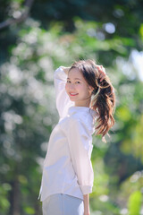 A young, smiling Asian woman in a white shirt and light pants, raising her hand to touch her ponytail, Posing outdoors in bright sun with a green bokeh background.